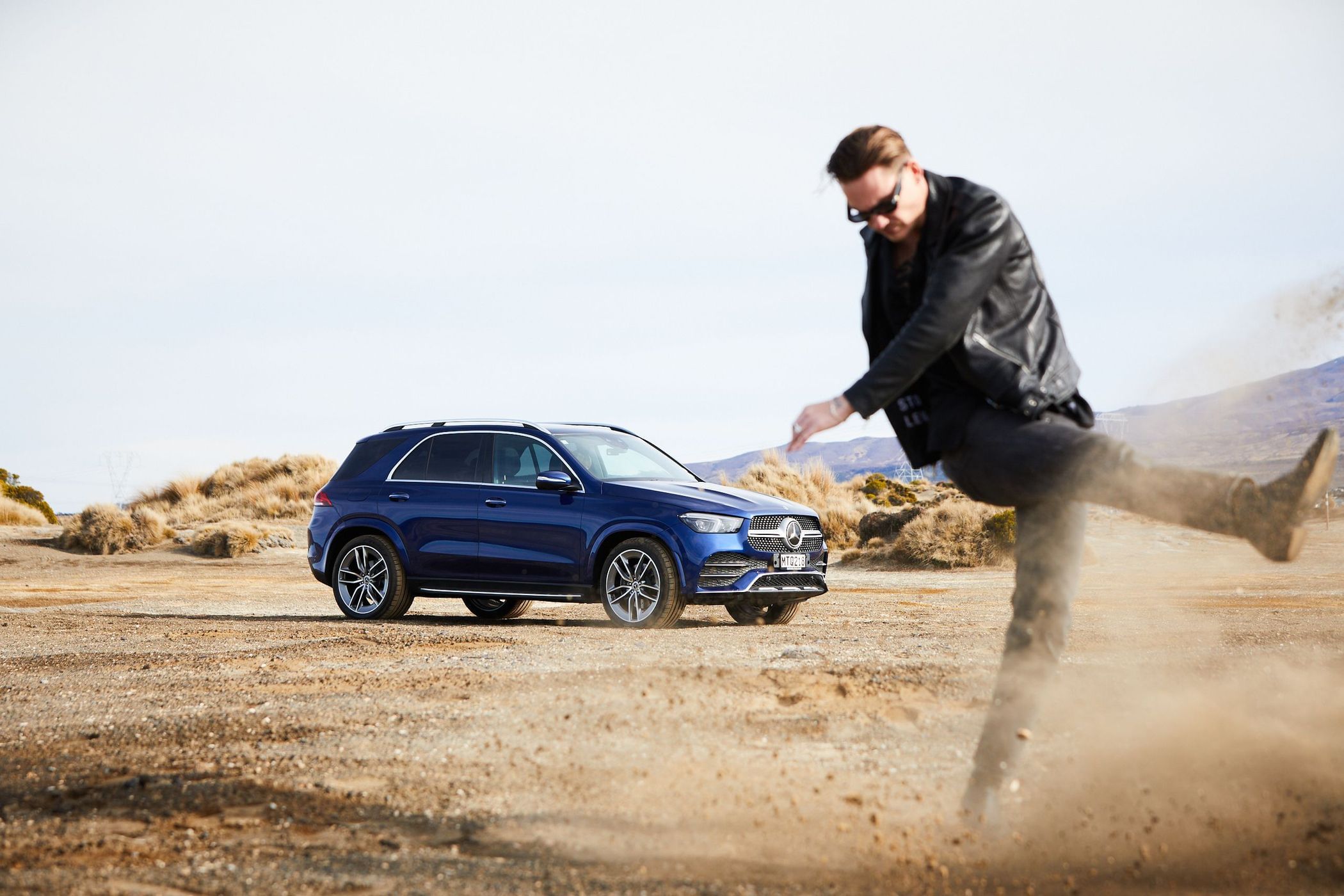 A blue Mercedes-Benz GLE SUV is parked on a desert landscape with a man kicking up sand in the foreground under clear skies.