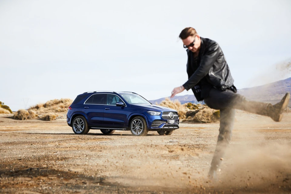 A blue Mercedes-Benz GLE SUV is parked on a desert landscape with a man kicking up sand in the foreground under clear skies.