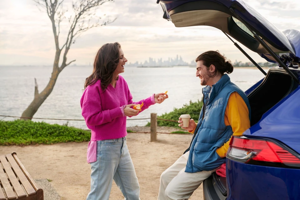 A woman shares an orange with a man by a blue car with an open trunk, near the ocean, under an overcast sky with a visible city skyline.