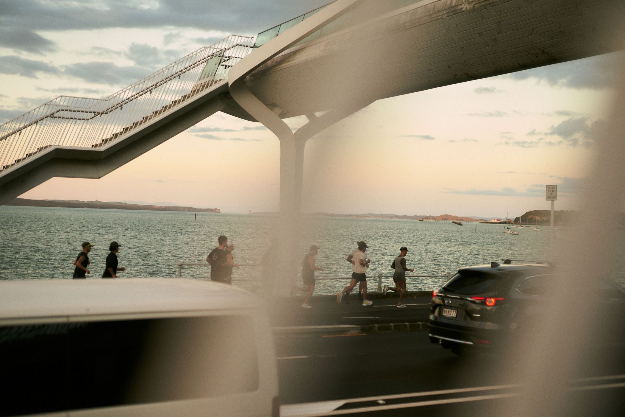 People jogging by the ocean under an overpass with cars on the street. Cloudy sky in the background. People running along the path near the water.