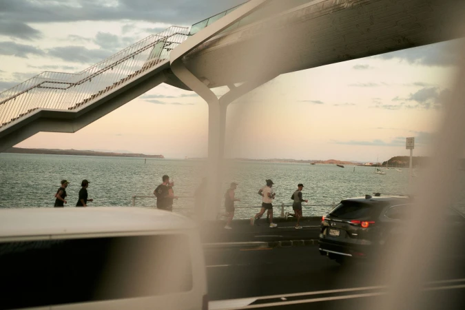 People jogging by the ocean under an overpass with cars on the street. Cloudy sky in the background. People running along the path near the water.