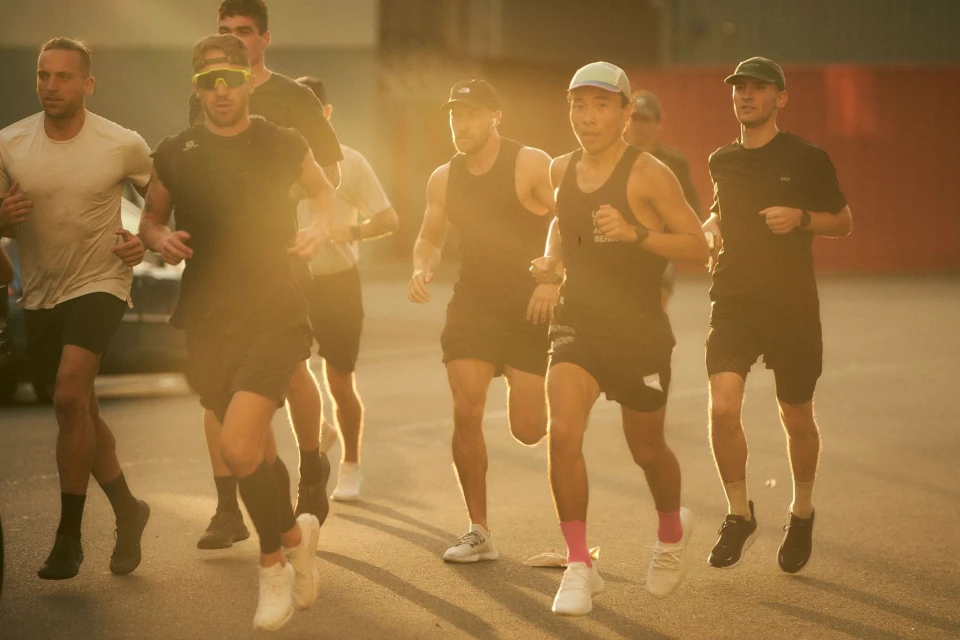 A group of people are running outside in athletic wear. Several adult men are moving quickly, wearing shorts and tank tops in the golden sunlight.
