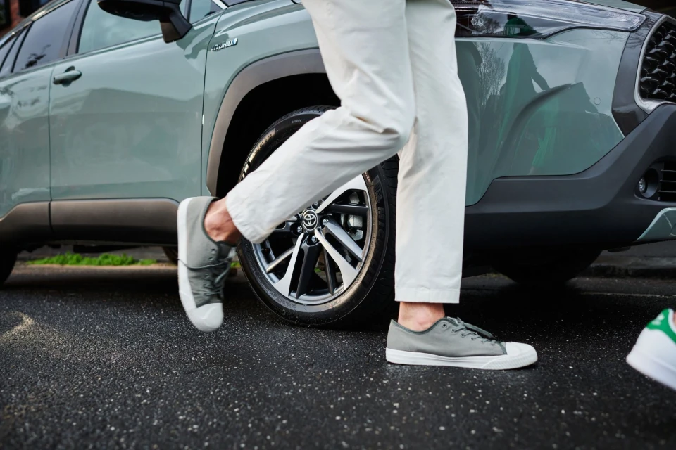 Close-up of a gray Toyota Corolla Cross Hybrid with a person wearing white pants and gray sneakers standing near the front tire.