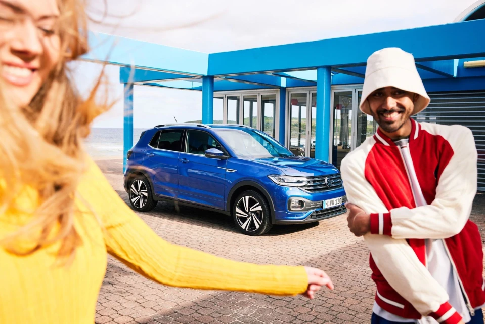 A blue Volkswagen T-Cross SUV is parked outdoors on a sunny day with two smiling young adults; automotive promotional image.