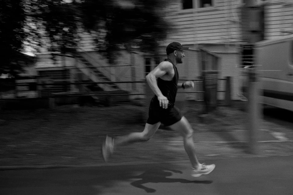 A man running outside at dusk wearing a cap and athletic wear, demonstrating speed with motion blur, on a road next to a house.