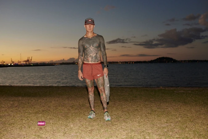 A tattooed man standing on a grassy shore wearing running shorts and a cap during sunset by the water.