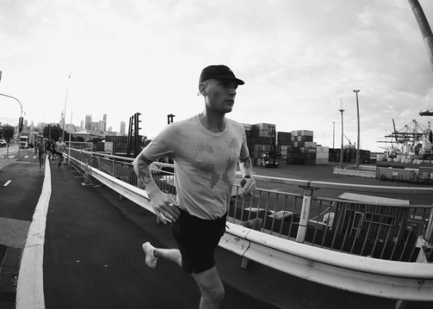 Man running on bridge with city skyline backdrop and shipping container area; black and white photo featuring a dynamic, active scene.