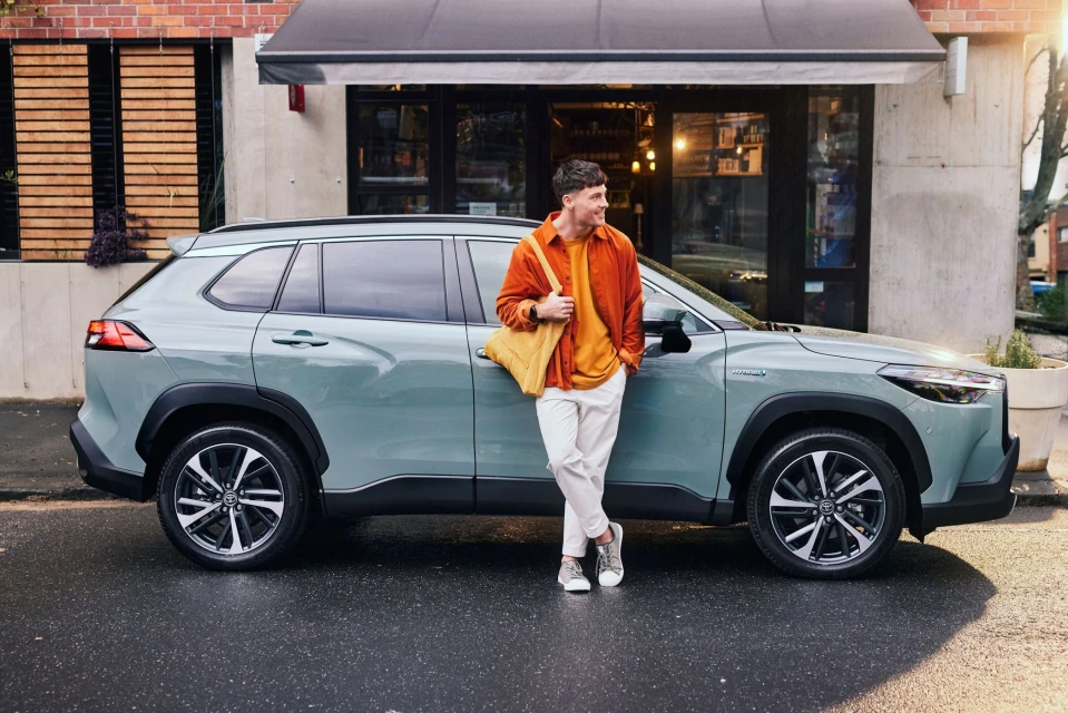 A light green Toyota Corolla Cross SUV parked on a wet street, with a man in an orange shirt leaning against the car. Urban background.