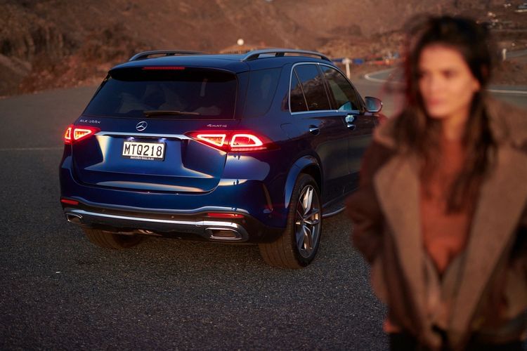 Dark blue Mercedes SUV parked on a paved road with an out-of-focus woman in the foreground, in a desert-mountain environment.