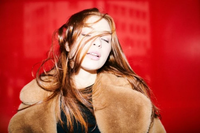 Close-up portrait of a woman with long red hair wearing a plush brown coat against a vibrant red background.