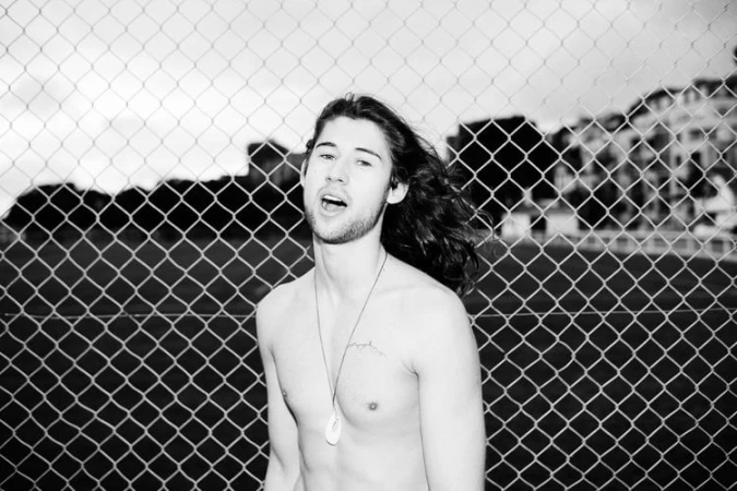 Monochrome portrait of a long-haired shirtless man wearing a pendant necklace against a chain-link fence by Bas&Chris.