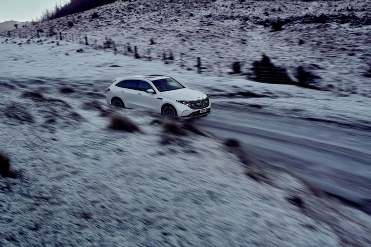 White Mercedes-Benz EQB SUV driving on a snow-covered road in a mountainous landscape. The scene evokes a sense of travel and adventure.