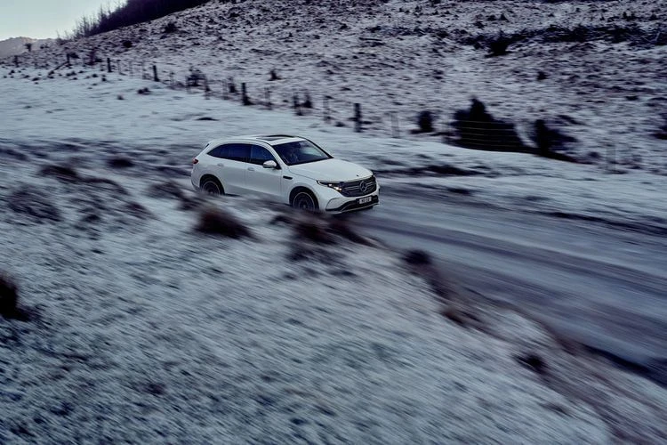 White Mercedes-Benz EQB SUV driving on a snow-covered road in a mountainous landscape. The scene evokes a sense of travel and adventure.
