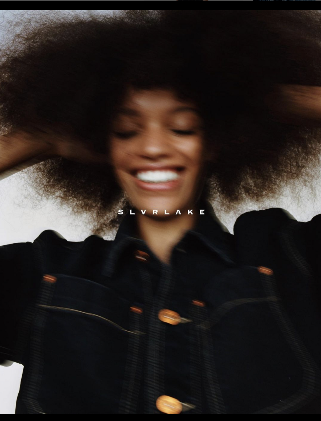 A smiling, dark-haired woman with an afro hairstyle wears a denim jacket in a blurry close-up shot with natural lighting.