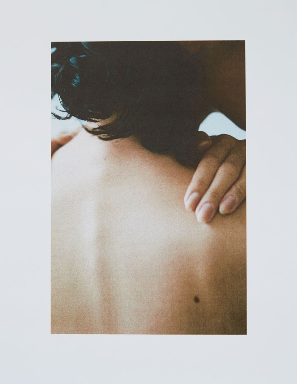 Close-up photograph of an adult's back with a hand resting on the shoulder, focusing on skin texture, a mole, and soft lighting.