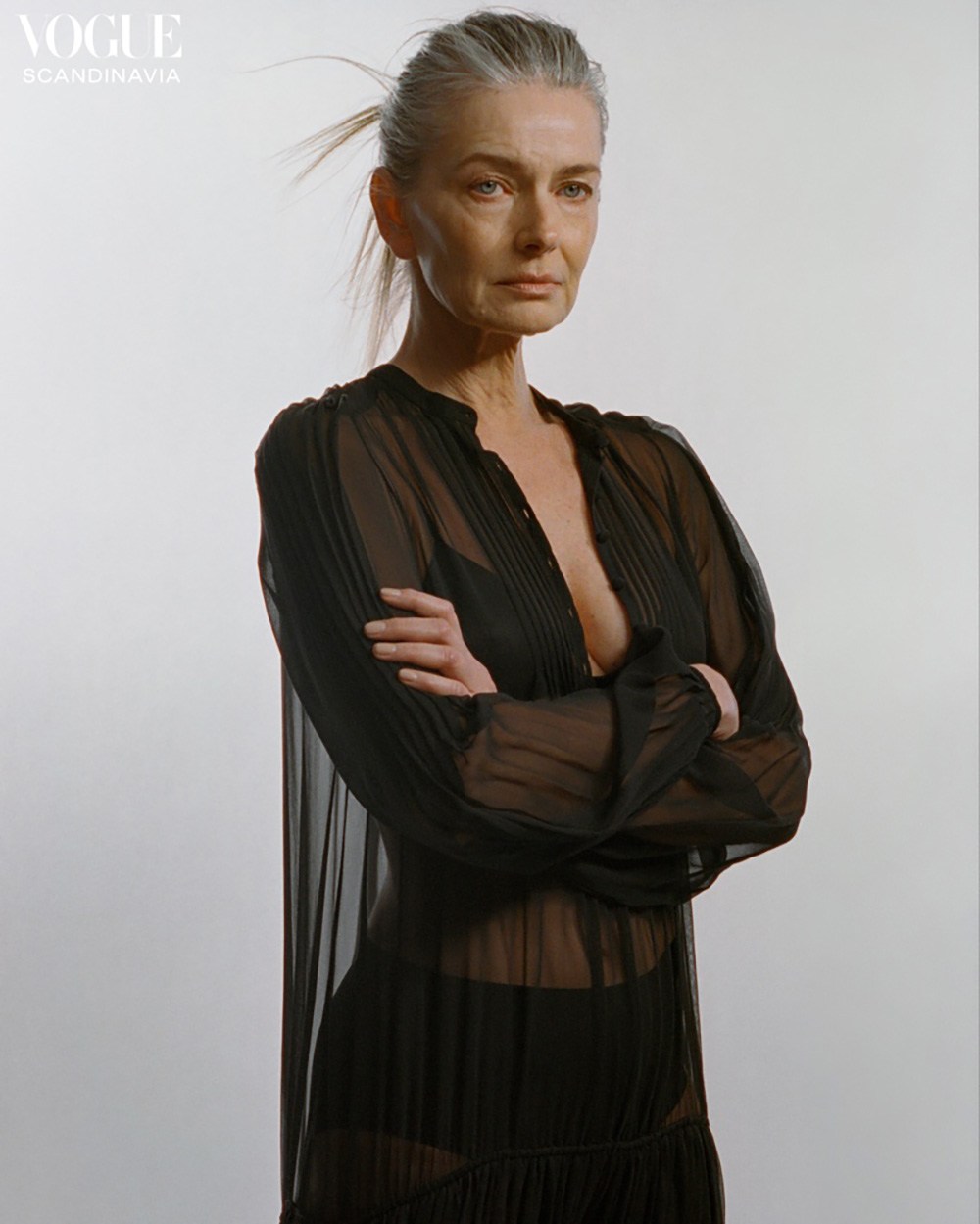 Mature woman with grey hair poses in a sheer black dress against a neutral background for a fashion editorial, arms crossed.
