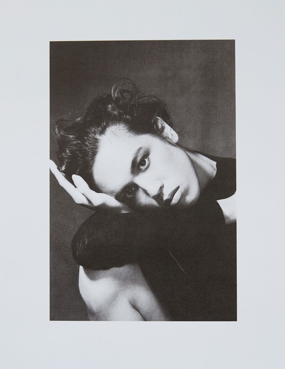 Portrait of a fair-skinned adult with curly hair, leaning on their arm against a dark background, with soft lighting and neutral expression.