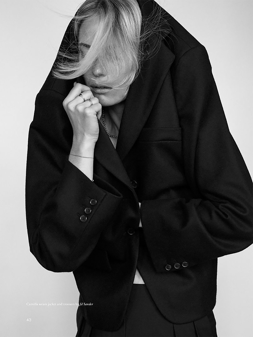 Black-and-white portrait of a woman with light blonde hair, wearing a black jacket over her head indoors with a white background.