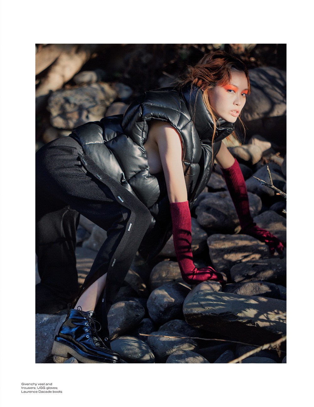 A young adult woman in a black puffer vest and knit gloves strikes a dynamic pose among the rocks and trees outdoors.