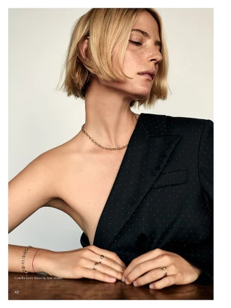 Model with blonde short hair wearing a black blazer and gold jewelry resting hands on a wooden table.