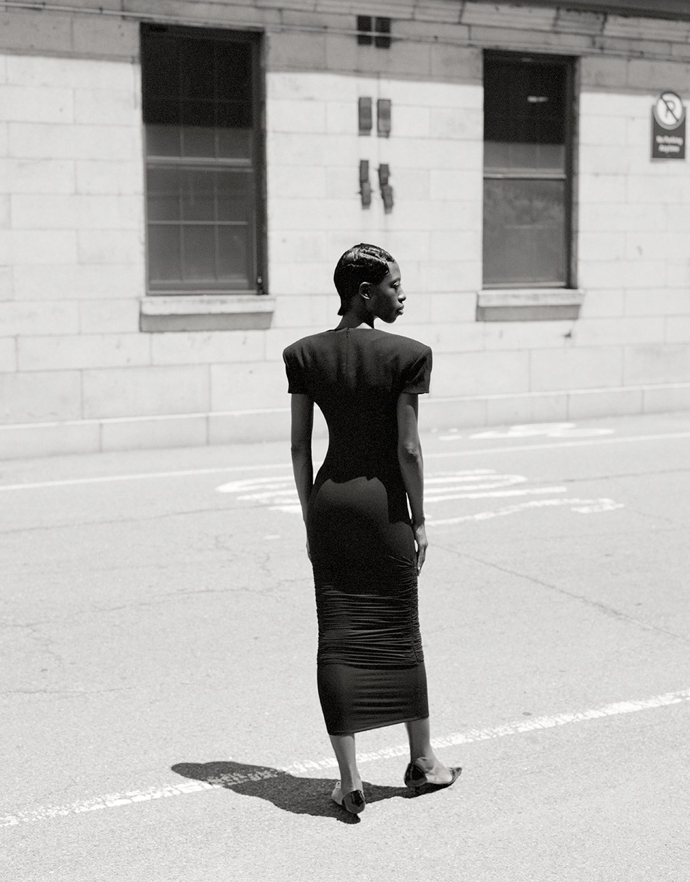 Elegant adult female in a black dress standing on a city street, showcasing fashion style and urban environment in black and white.