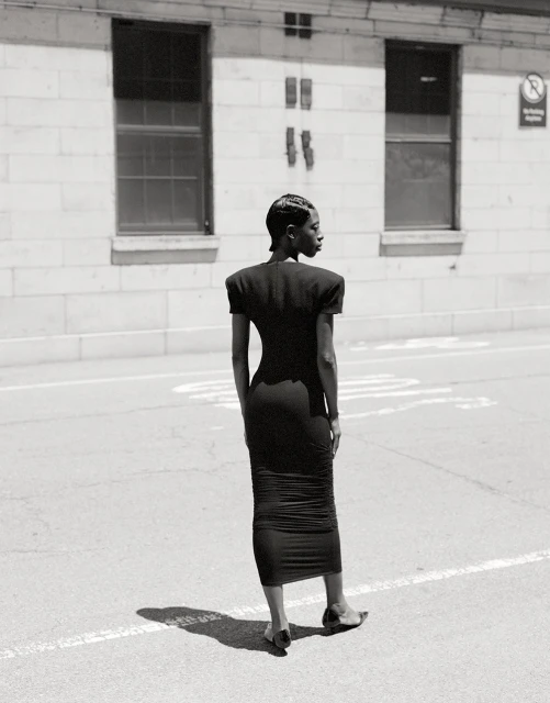 Elegant adult female in a black dress standing on a city street, showcasing fashion style and urban environment in black and white.