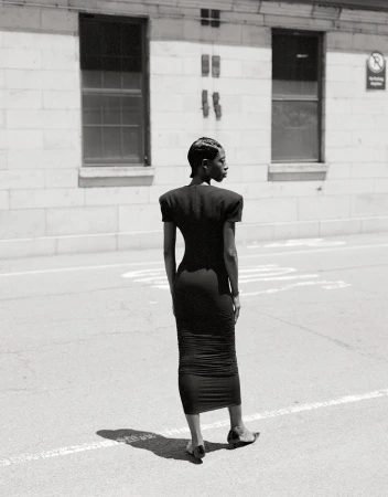 Elegant adult female in a black dress standing on a city street, showcasing fashion style and urban environment in black and white.