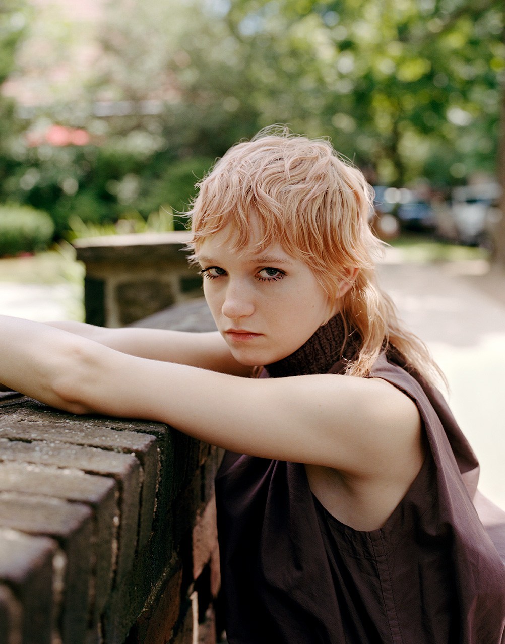 Portrait of a young woman with wavy reddish-blonde hair leaning against a brick structure in an outdoor setting with a blurred background.