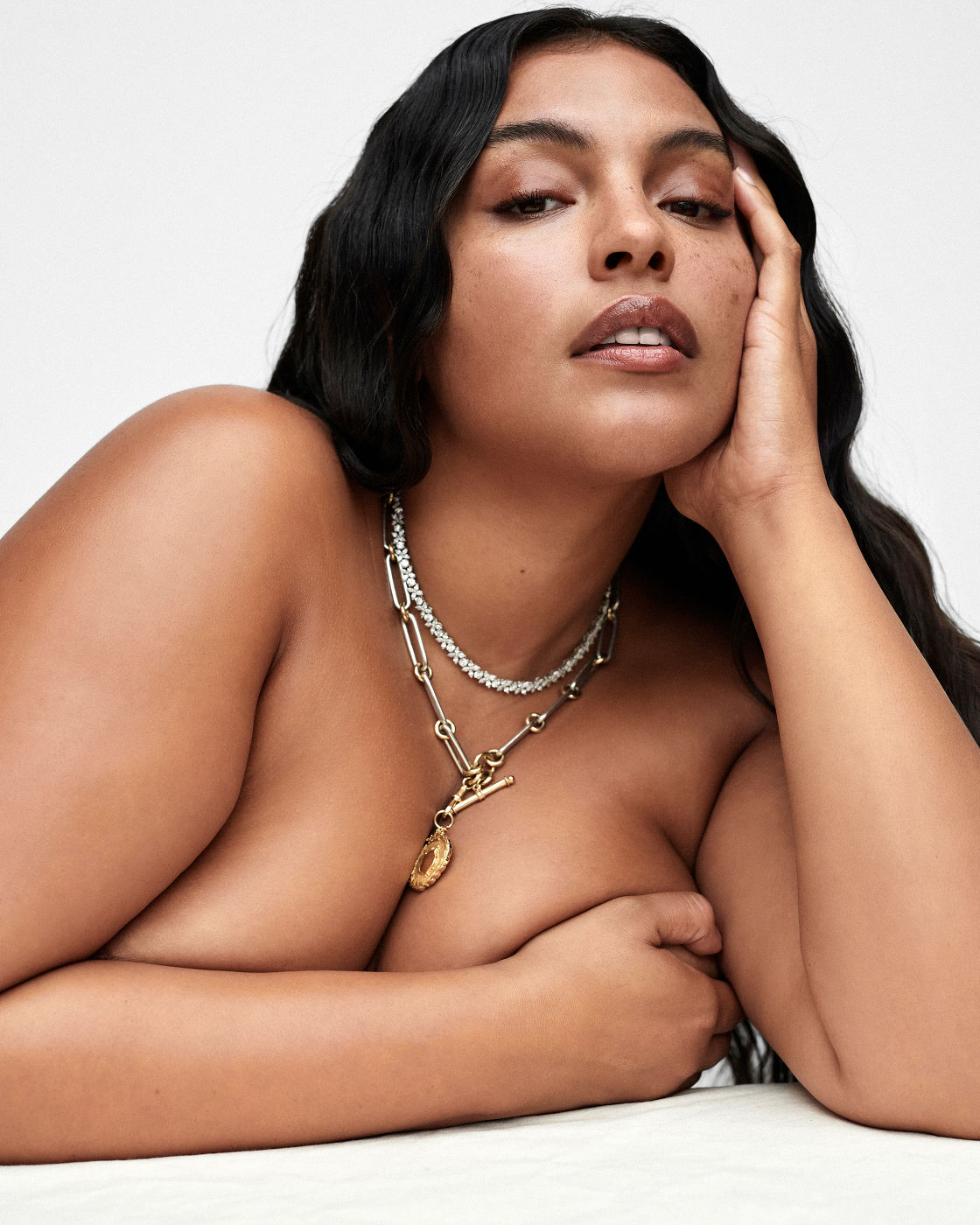 Portrait of a woman wearing layered necklaces with her hand to her face against a white seamless background