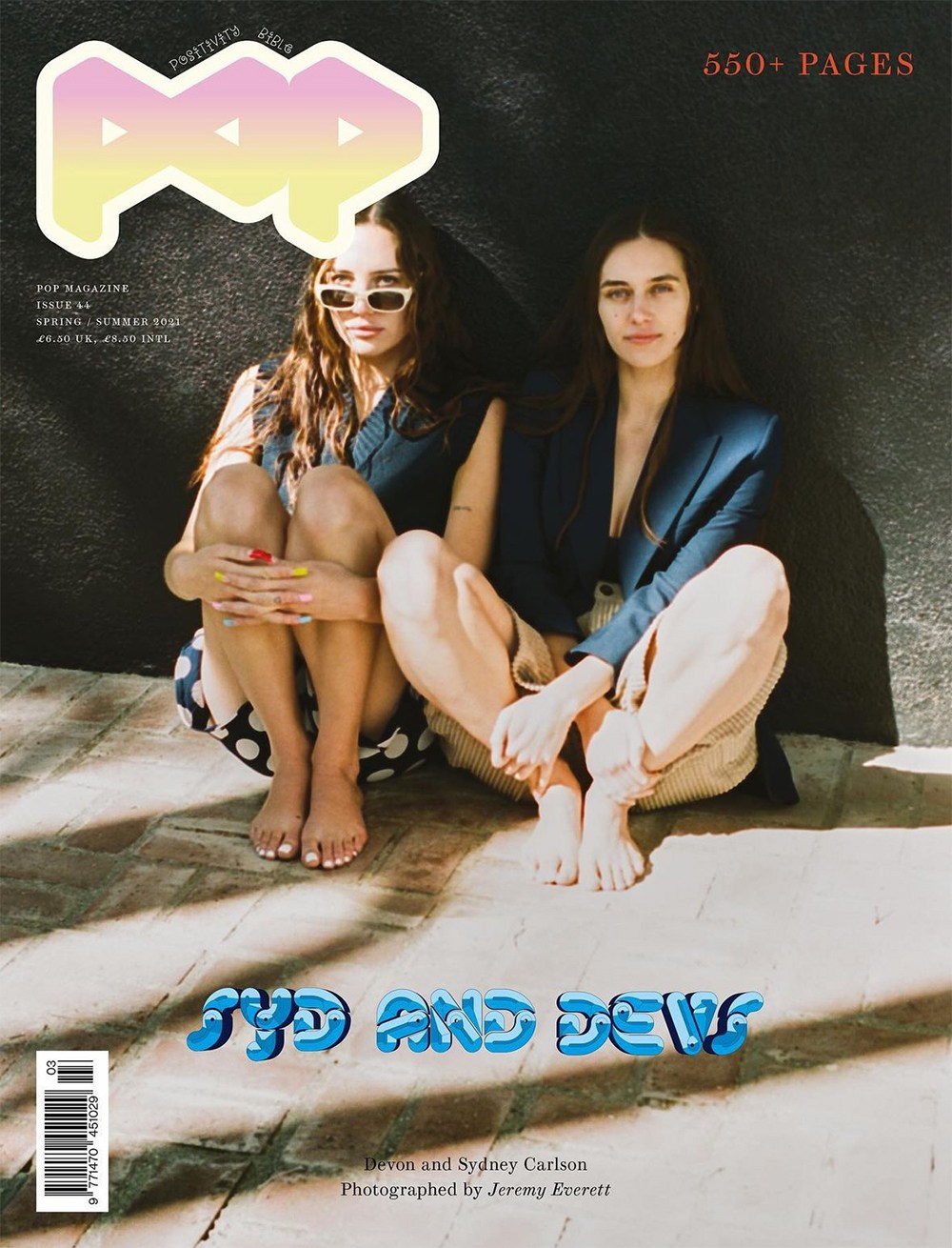 Two adult women pose seated on a brick surface in a fashion magazine cover, wearing dark blazers, neutral pants, in a natural outdoor light.