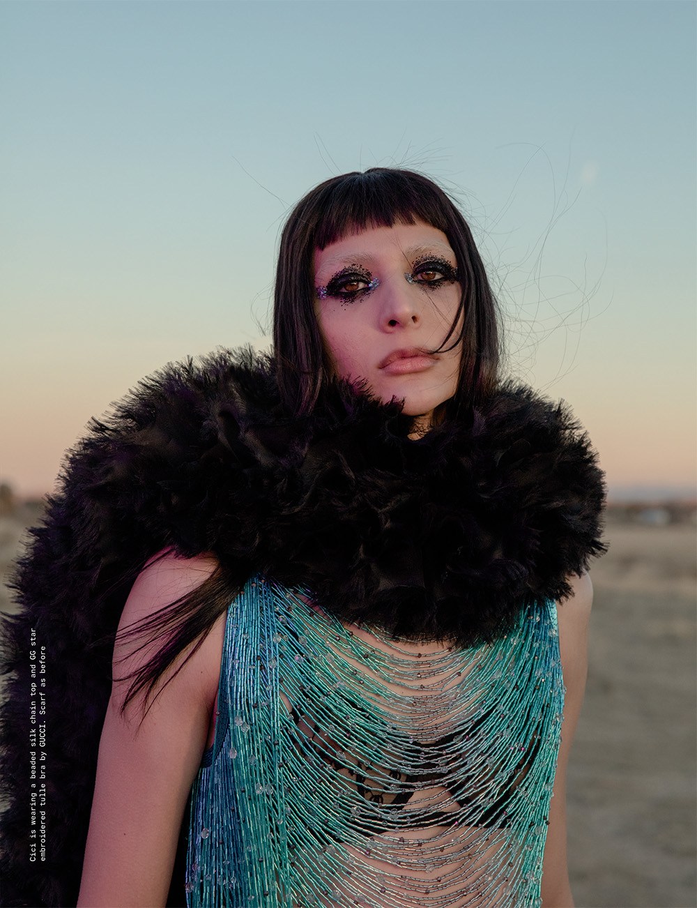 A woman with dramatic makeup and a black feathered shawl poses in a shimmering gown against a soft twilight desert backdrop.
