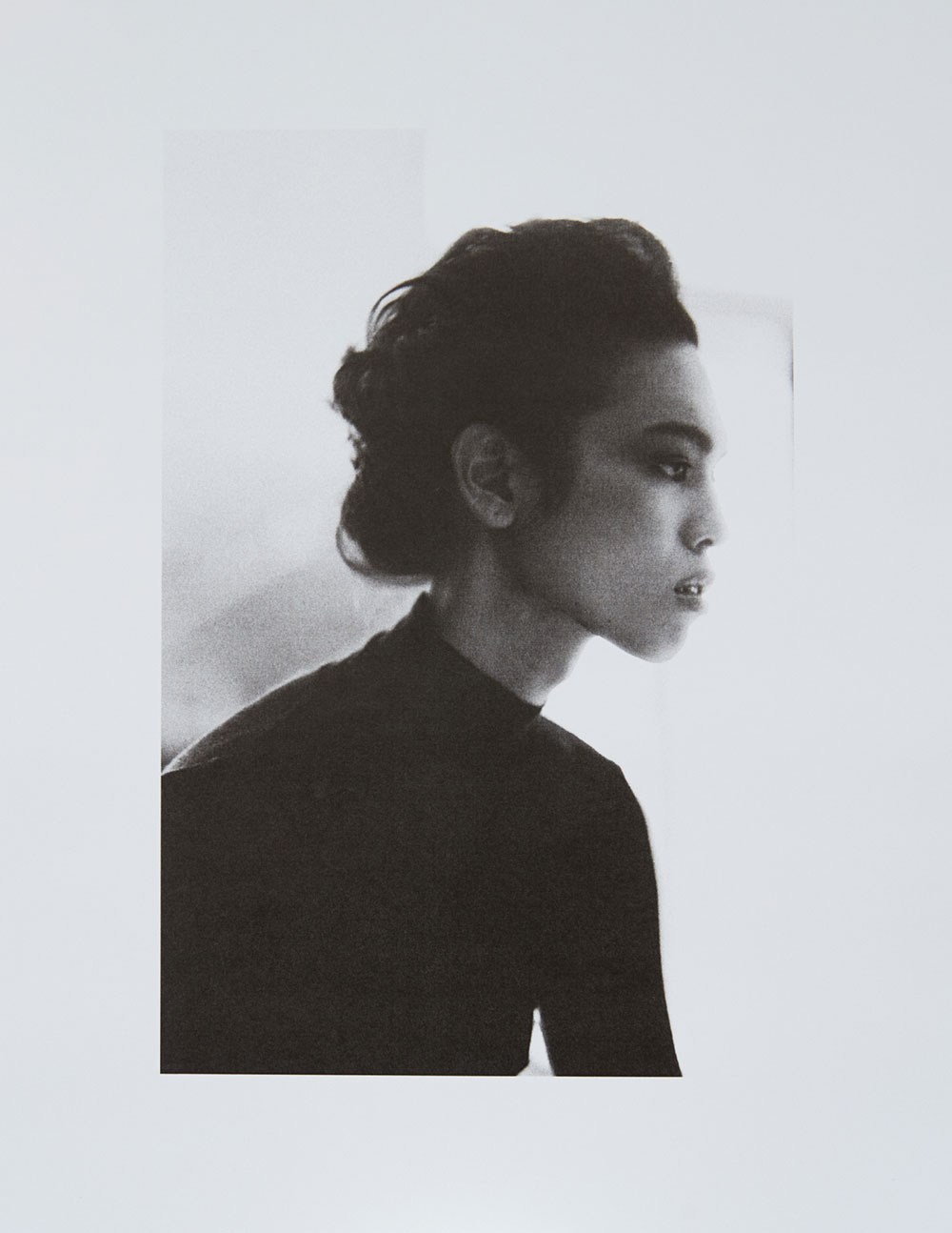 Monochromatic portrait of a young adult woman in profile, wearing a black top and styled hair, against a soft white background.