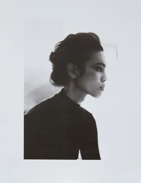 Monochromatic portrait of a young adult woman in profile, wearing a black top and styled hair, against a soft white background.