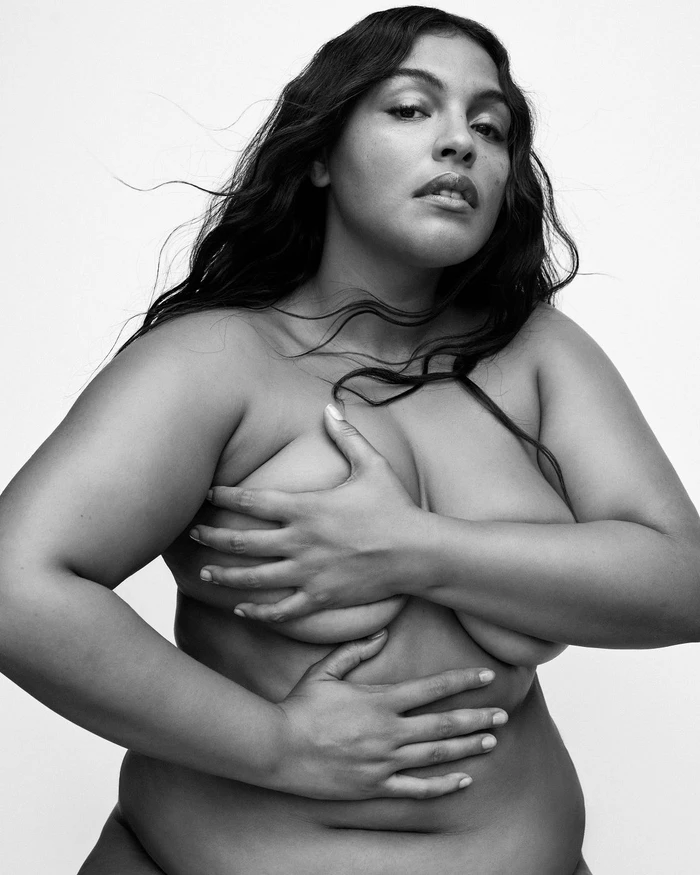 Black and white portrait of a woman with long dark wavy hair holding her hands across her chest against a plain background