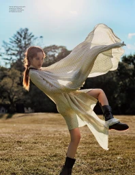 Model in flowing patterned dress and dark boots kicking leg outward in park under sunny sky by Jenny Kim