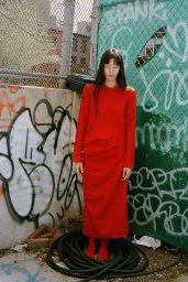 Model in long red dress standing outdoors against a graffiti-covered wall with chain-link fence in New York by Jenny Kim
