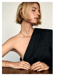 Model with blonde short hair wearing a black blazer and gold jewelry resting hands on a wooden table.