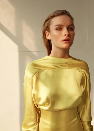 Close-up portrait of a woman in a glossy yellow satin top with sunlight casting diagonal shadows across a light wall