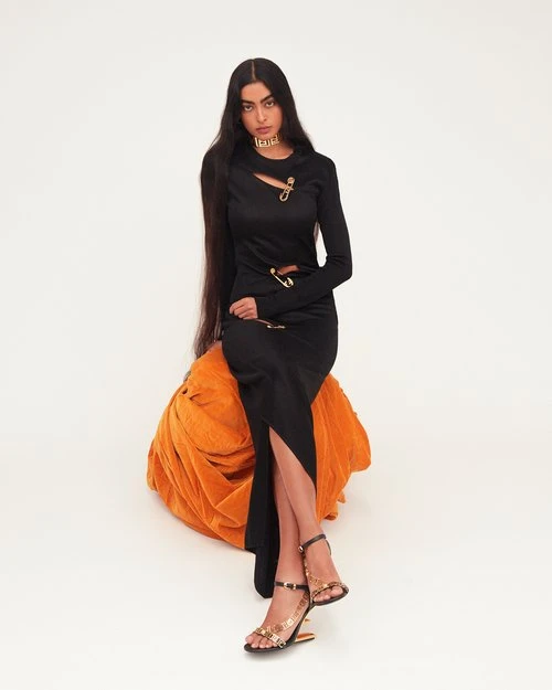 Fashion portrait of a woman with long dark hair, wearing a black dress and gold heels, sitting on an orange plush seat against a white backdrop.