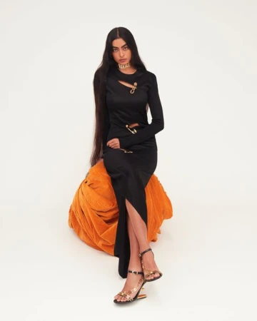 Fashion portrait of a woman with long dark hair, wearing a black dress and gold heels, sitting on an orange plush seat against a white backdrop.