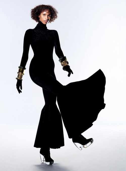 Fashion editorial shot of a Black woman in a black jumpsuit and clear platform heels strikes a pose against a white backdrop. Gold bracelets.