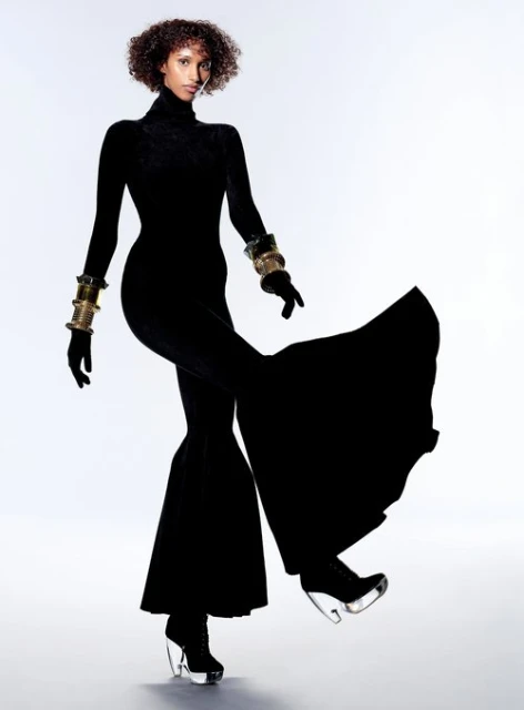 Fashion editorial shot of a Black woman in a black jumpsuit and clear platform heels strikes a pose against a white backdrop. Gold bracelets.