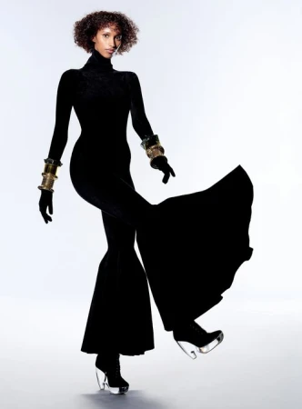 Fashion editorial shot of a Black woman in a black jumpsuit and clear platform heels strikes a pose against a white backdrop. Gold bracelets.