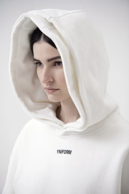 Close-up of a young woman wearing a white hooded sweatshirt with the YNIFORM logo. Shot in a bright, neutral photo studio.
