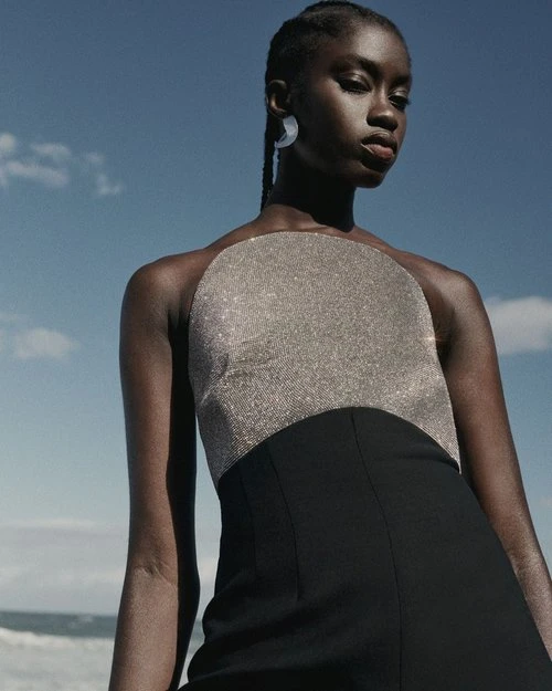 Elegant portrait of a Black woman outdoors, wearing a silver and black halter dress against a soft blue sky and distant ocean.