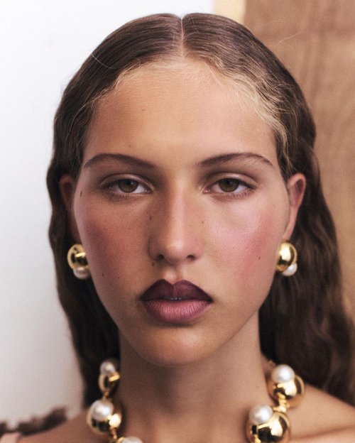 Close-up studio shot of a young adult female with dark hair, statement gold jewelry, and deep plum lipstick against a neutral backdrop.