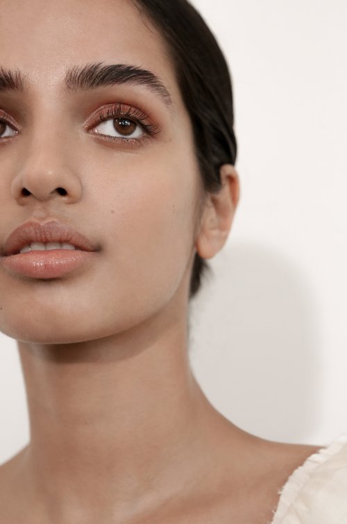 Close-up portrait of a woman with brown eyes and dark hair, featuring natural makeup and soft, diffused lighting against a light background.
