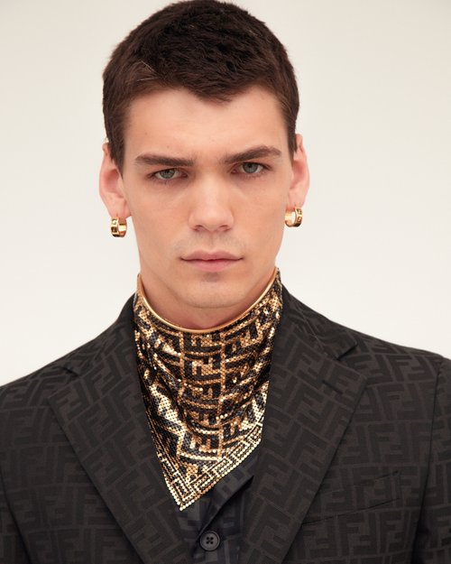 Portrait of a man wearing a patterned black suit, high-collared patterned neckpiece, and small gold hoop earrings.