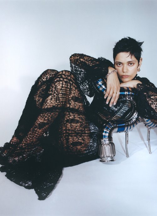 A woman with short dark hair poses in a black lace dress while leaning on a chrome object in a studio setting.