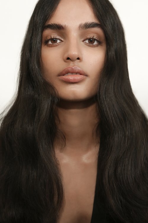 Close-up portrait of an adult woman with long, dark wavy hair against a plain white background, featuring a direct gaze and soft, neutral makeup.